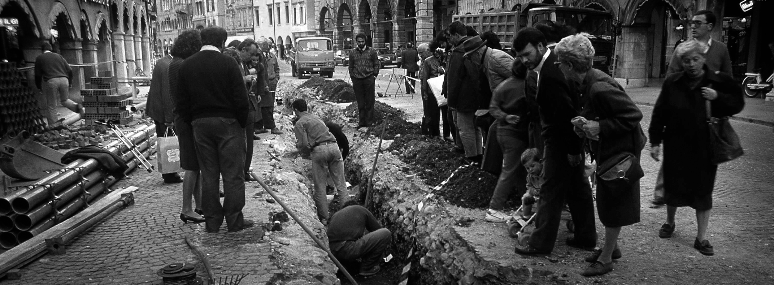 Fotografia scattata negli anni '80 in occasione delle indagini archeologiche in via Mercatovecchio a Udine che ritrae alcuni cittadini curiosi e gli archeologi impegnati nell'attività di scavo.